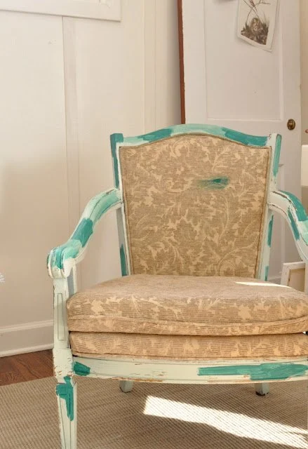 Embellishing an Upholstered Chair with Doilies 11 An upholstered armchair with a beige patterned fabric and a distressed wooden frame, featuring areas of mint green and turquoise paint, sits on a light-colored rug.