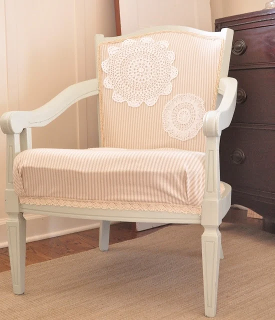 Embellishing an Upholstered Chair with Doilies 12 A vintage armchair with a white painted frame and beige striped upholstery, adorned with lace doilies on the backrest, stands on a woven rug near a dark wooden dresser, showcasing the charm of furniture makeovers in its renewed elegance.