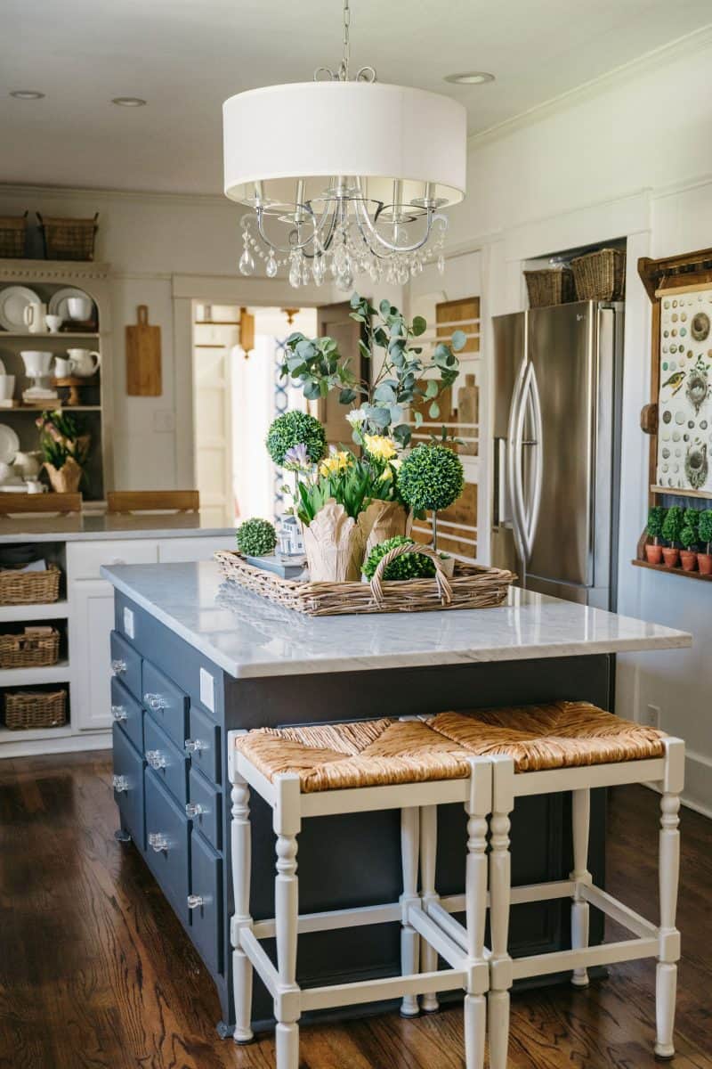 a cozy and elegant kitchen featuring a blue island with a marble countertop, white wooden stools, a chic pendant light, and open shelving stocked with dishes in the background.