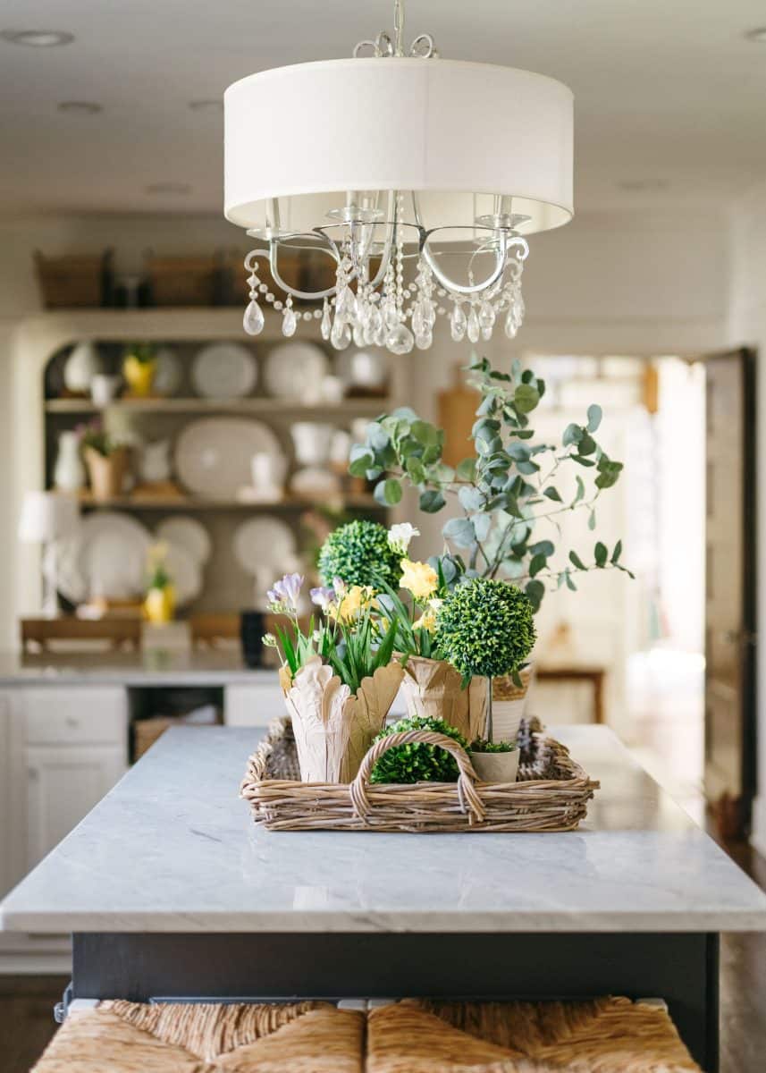 a cozy interior scene with a rustic wooden table topped with a marble surface, elegantly decorated with a woven basket containing an assortment of green plants and flowers adding a touch of nature, under the warm glow of a classic chandelier with crystal accents.