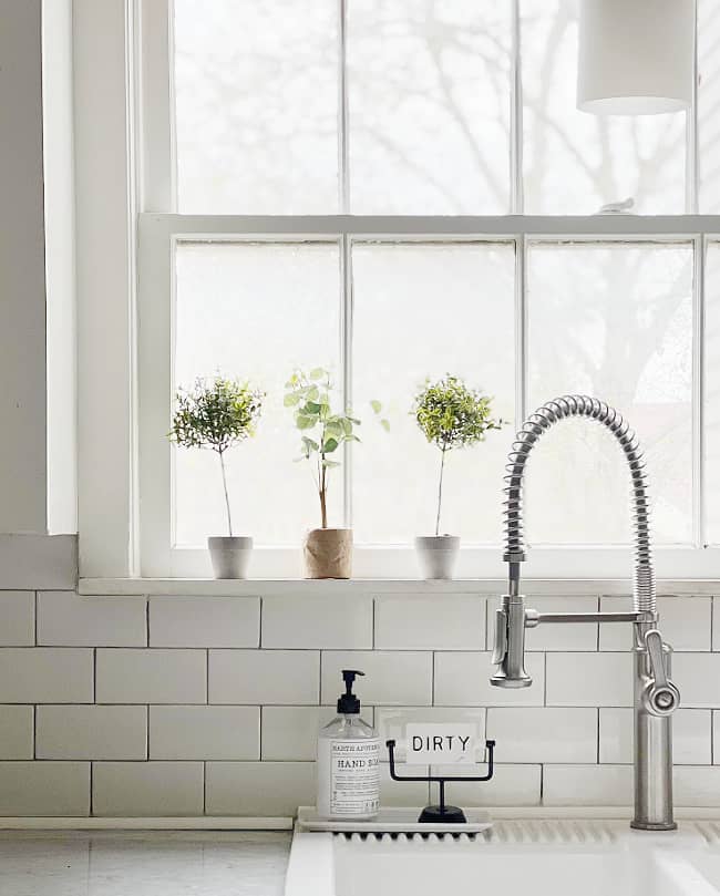 a serene kitchen scene with a clean, white aesthetic, featuring a modern faucet and three potted plants basking in the natural light from a large window on the kitchen island.