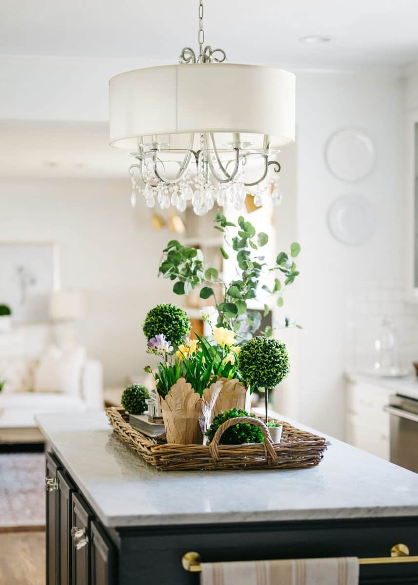 elegant kitchen interior featuring a stylish chandelier above a kitchen island with a decorative wicker basket filled with fresh green plants and flowers.
