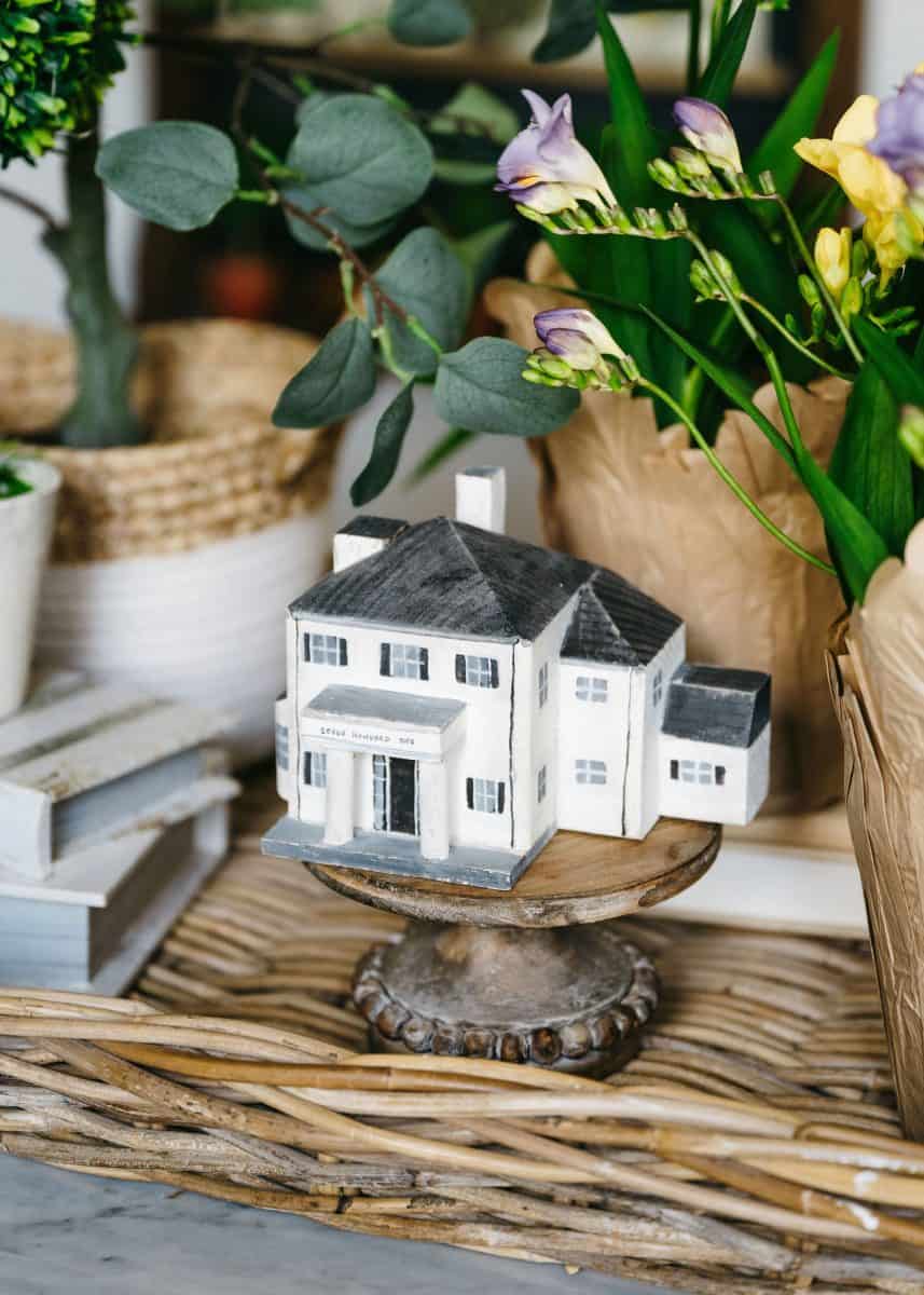 a miniature model of a white house displayed on a rustic wooden stand, surrounded by fresh green plants and vibrant flowers, creating a cozy and charming home decor vignette.