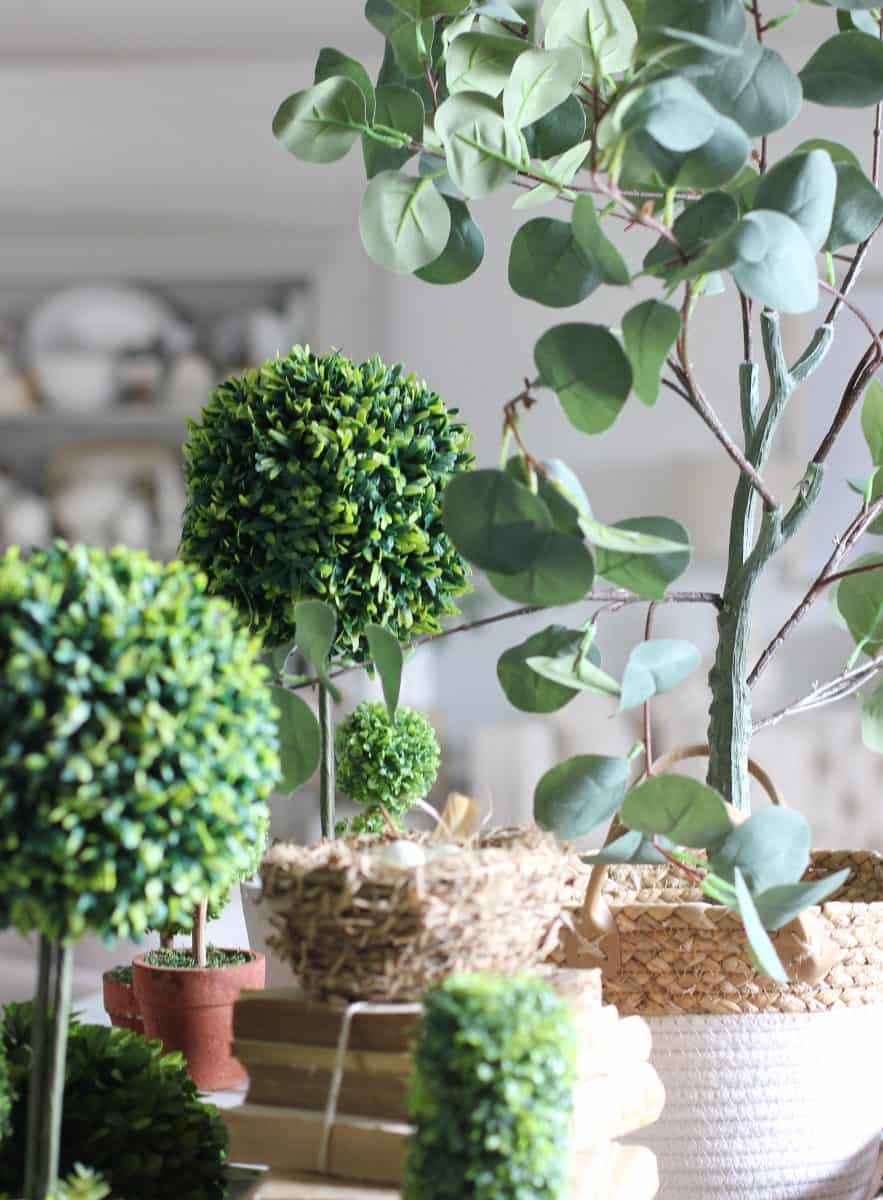 a collection of green artificial topiary plants and a eucalyptus in various pots and baskets, arranged to create an indoor decorative garden setting on a kitchen island.