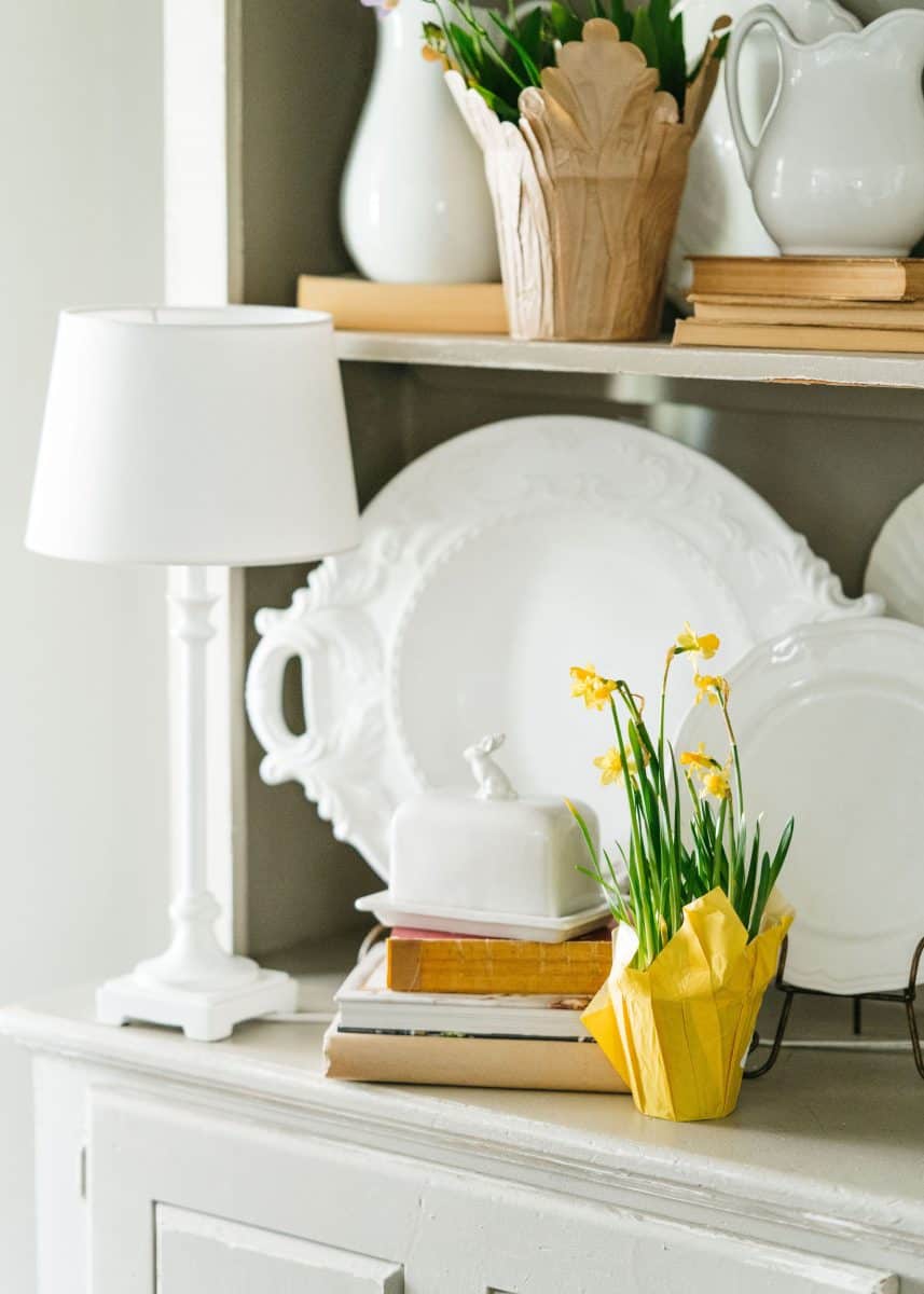 a neatly arranged home interior with a collection of decorative items, including a white table lamp, porcelain dishware, a small potted plant with yellow flowers, and a stack of books arranged on a white-painted wooden hutch.
