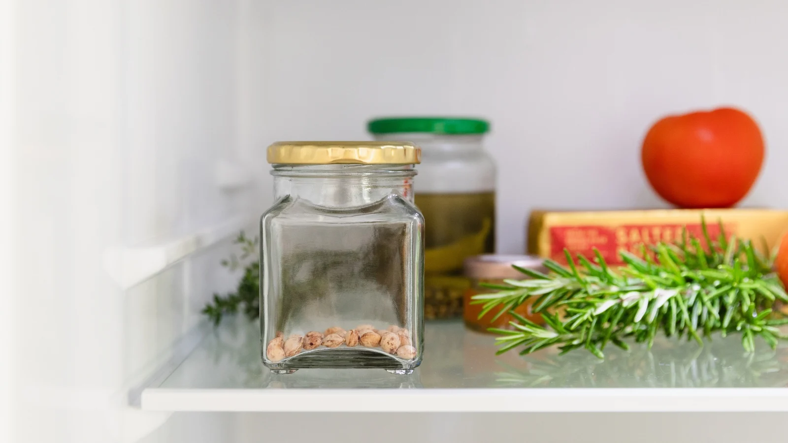 a small glass jar with a metal lid containing small round ovules, placed on a white shelf on a refrigerator, next to rosemary and a red fruit.