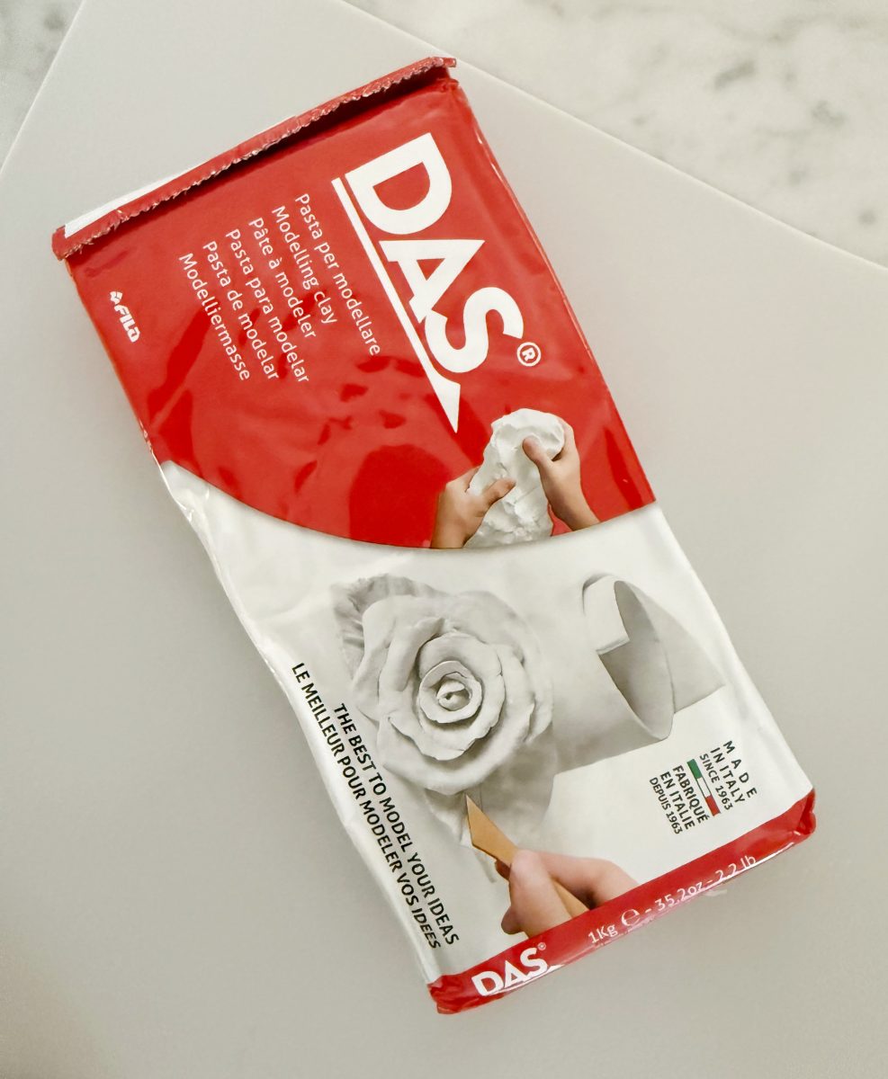 a package of das modeling clay, mostly white with a red section showing the brand name. the packaging features images of hands sculpting a clay rose and shaping a piece of clay.