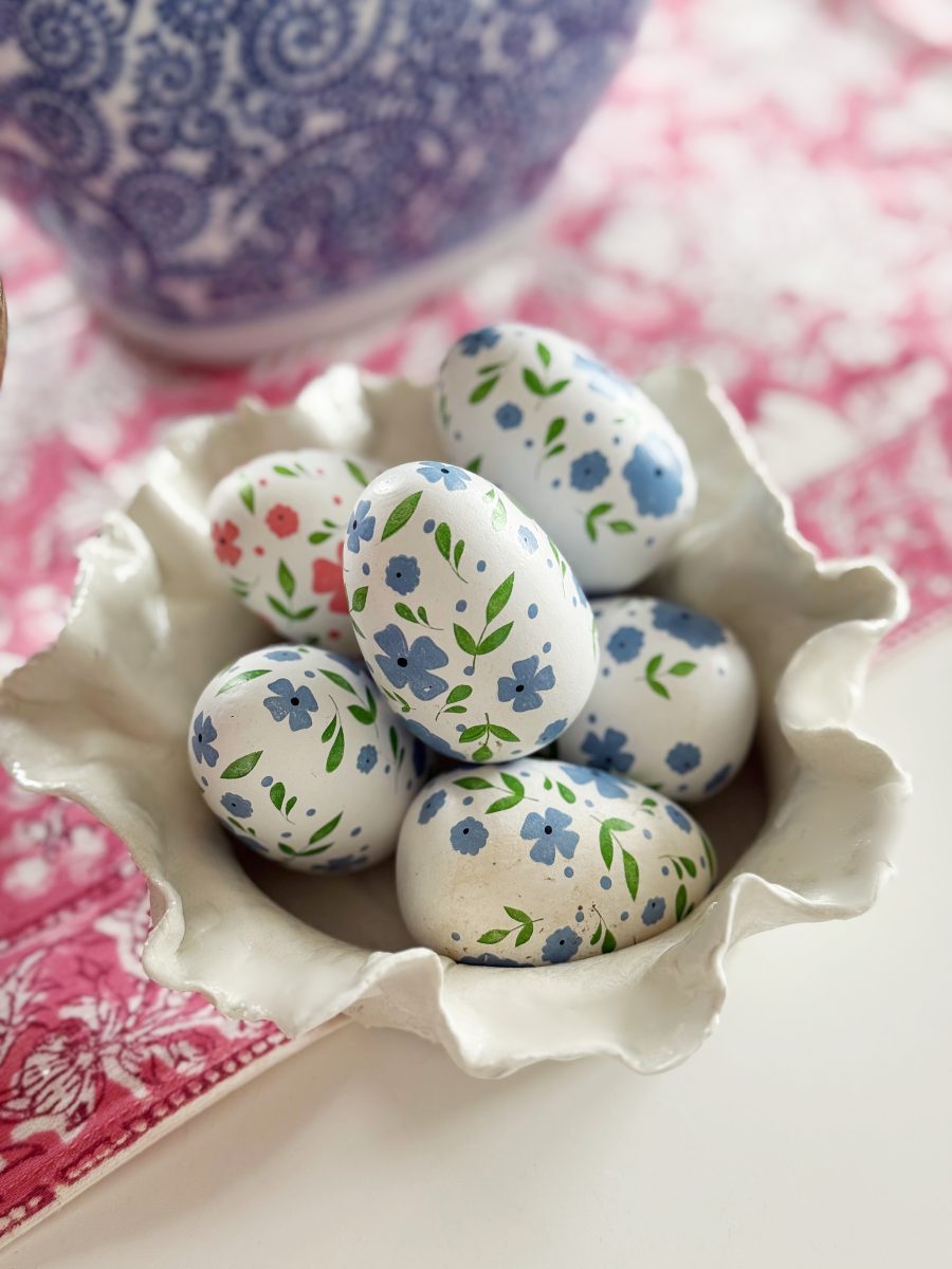 a ceramic bowl holds several white eggs decorated with hand-painted blue, green, and red floral patterns, placed on a pink patterned cloth with a blue and white vase in the background.