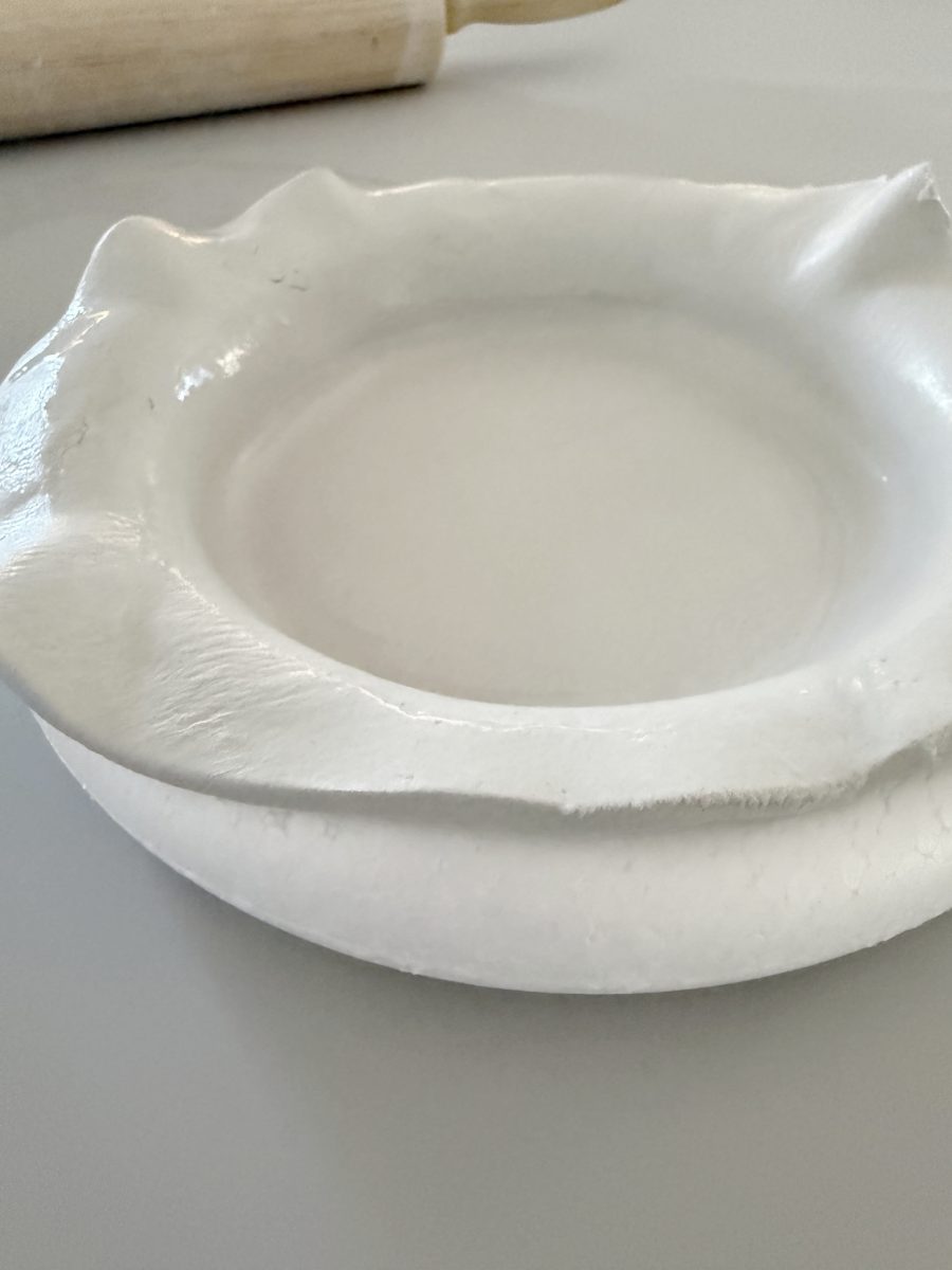 a pie dish with a sheet of uncooked pie dough draped over the edges sits on a counter. a wooden rolling pin is partially visible in the background.