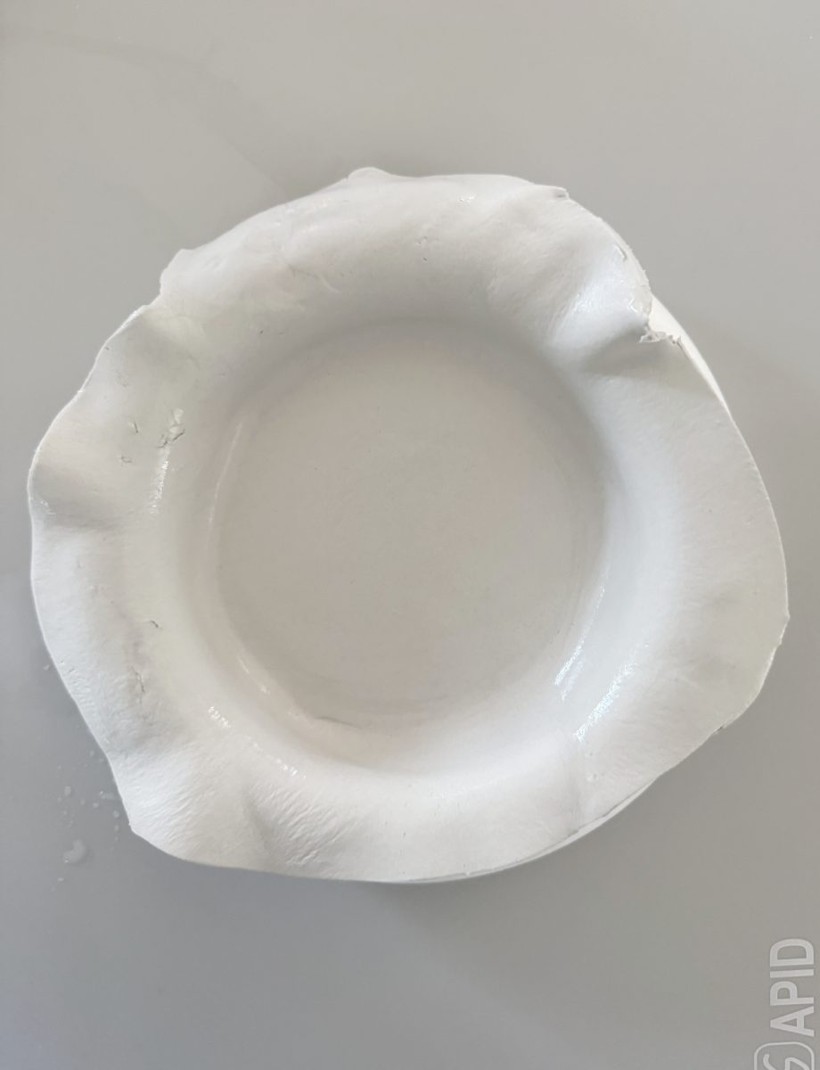 a flat, round disc of white clay lies on a light gray surface. the clay has subtle cracks and textured lines on its surface, with uneven edges and a small piece protruding from the bottom.
