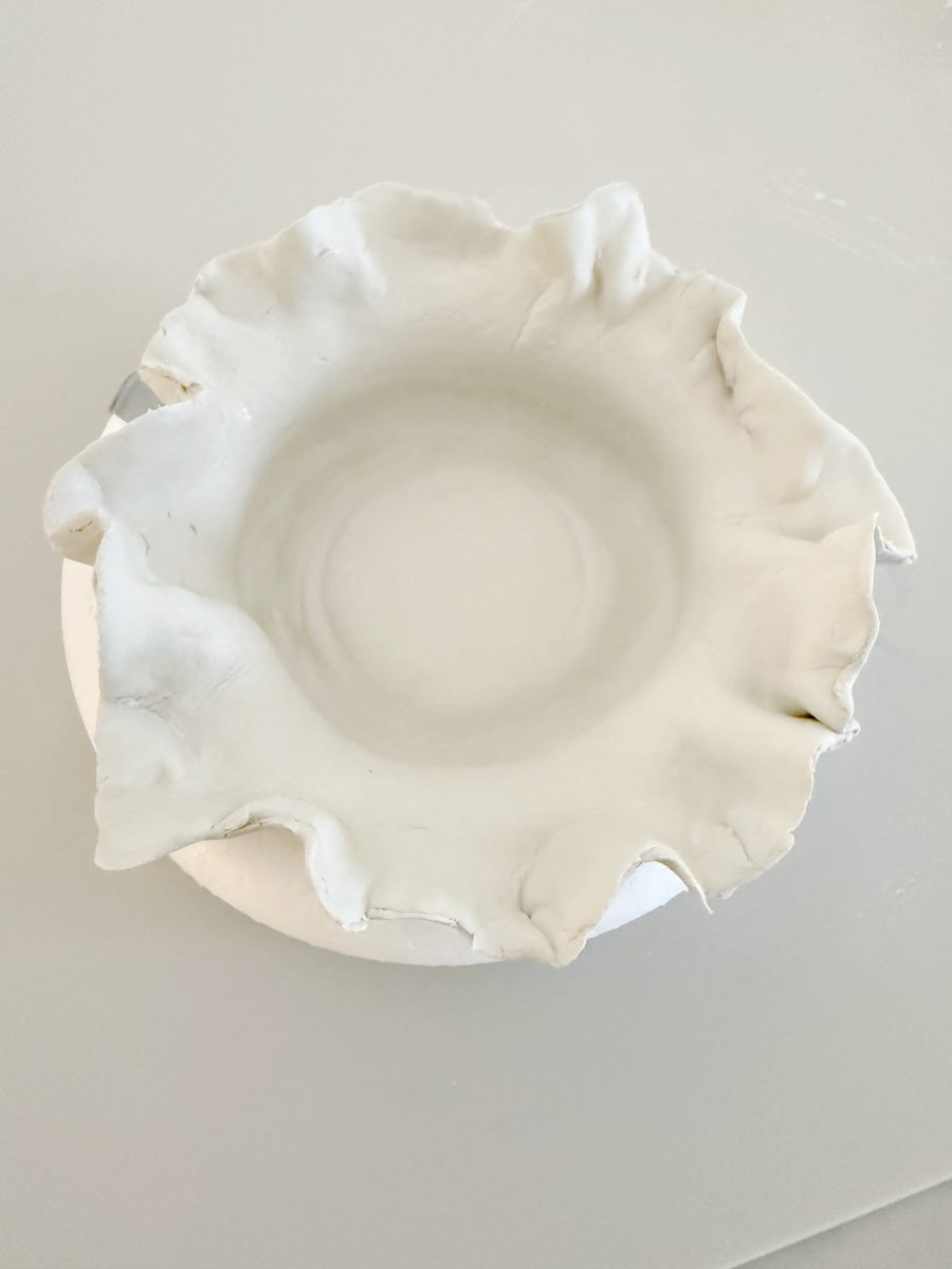 a white ceramic bowl with an irregular, wavy rim sits on a smooth, light-colored surface. the bowl has a handmade, organic appearance.