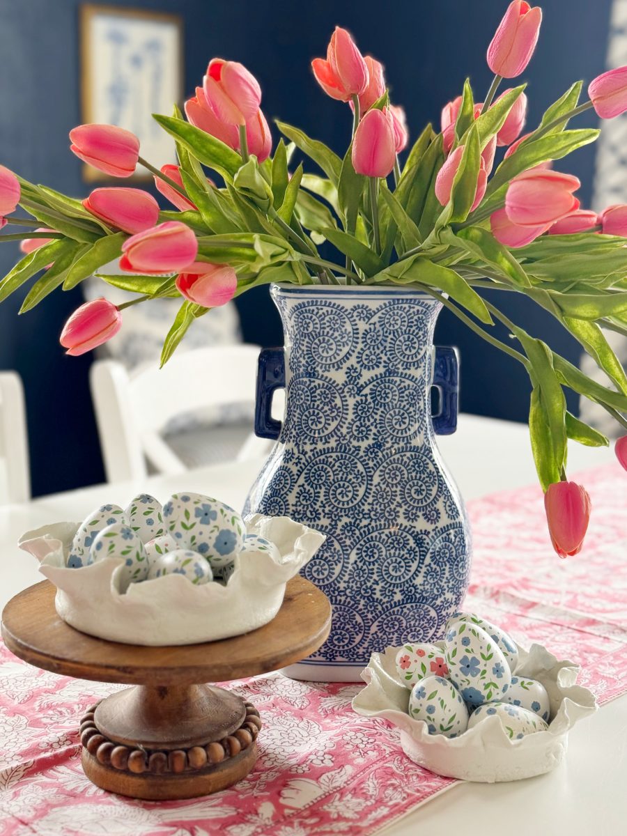 a blue and white vase with pink tulips sits on a table with a pink runner. decorative eggs with floral patterns are displayed in two white bowls, one elevated on a wooden stand.