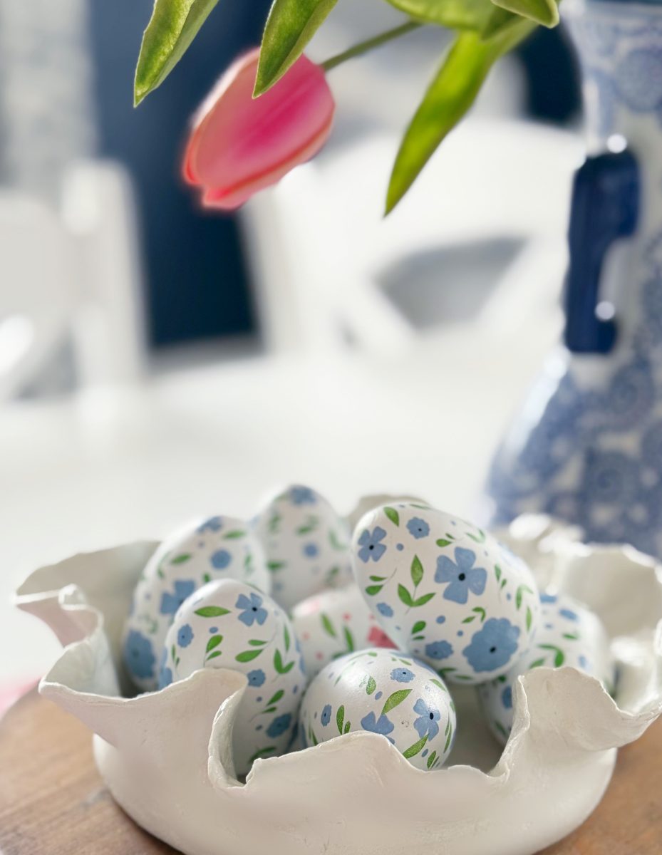 a white ceramic bowl holds painted eggs with blue floral patterns. a pink tulip and part of a blue-patterned vase are visible in the soft-focus background.