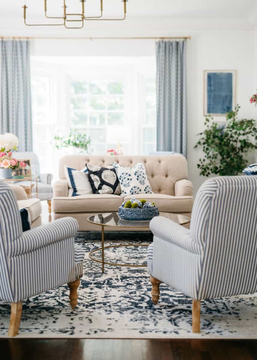 how to mix patterns with blue and white 2 bright living room with striped armchairs, a beige tufted sofa, patterned cushions, round glass coffee table with a bowl of apples, blue and white rug, large windows, blue curtains, and indoor plants.