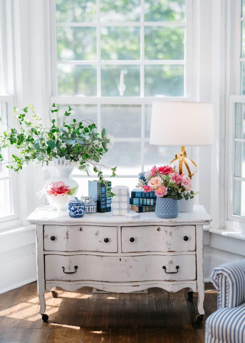 how to mix patterns with blue and white 1 a white vintage dresser sits by large windows, topped with books, a gold lamp, a vase of pink flowers, greenery in a white vase, and blue-and-white ceramics, in a sunlit, airy room with wood floors.