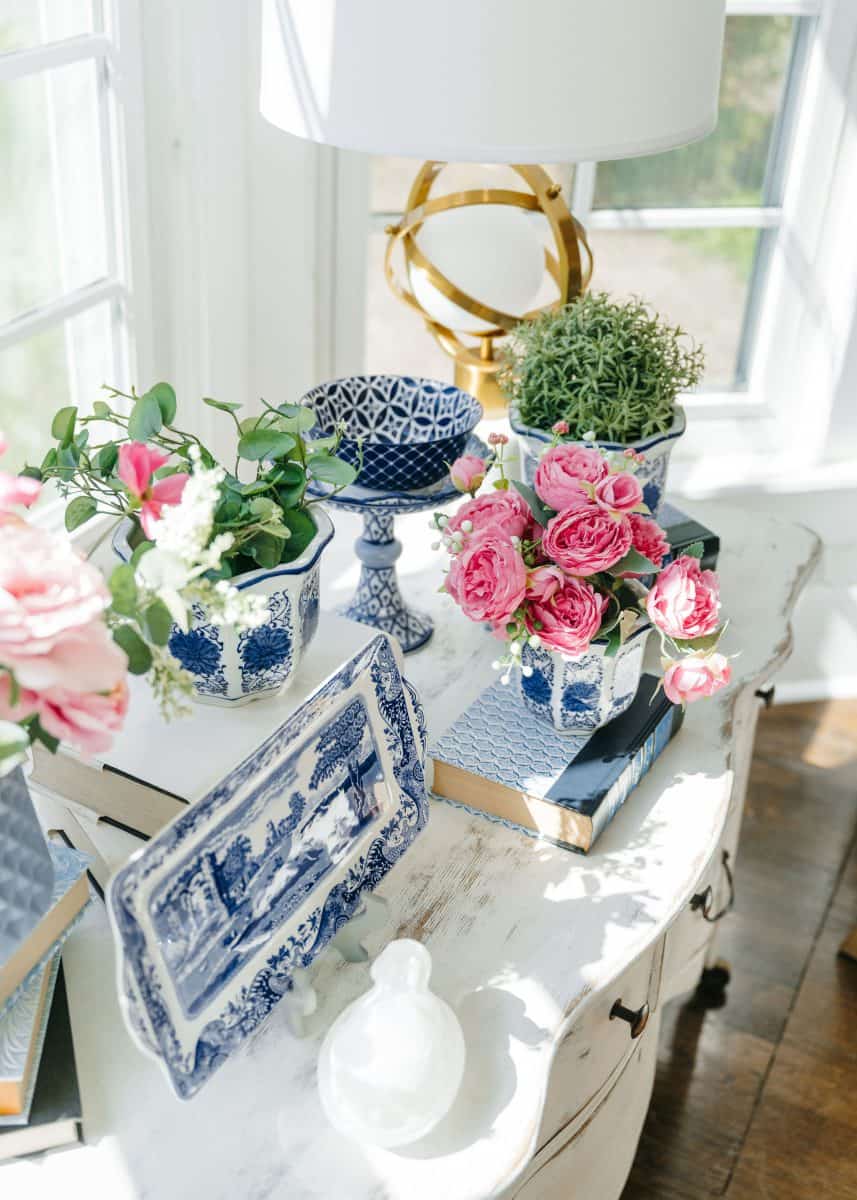 how to mix patterns with blue and white 14 a cozy corner featuring a vintage white table adorned with a classic blue and white porcelain vase filled with pink roses, a stack of books, a small potted plant, and an elegant table lamp with a golden base and a white lampshade casting a warm glow by the window.