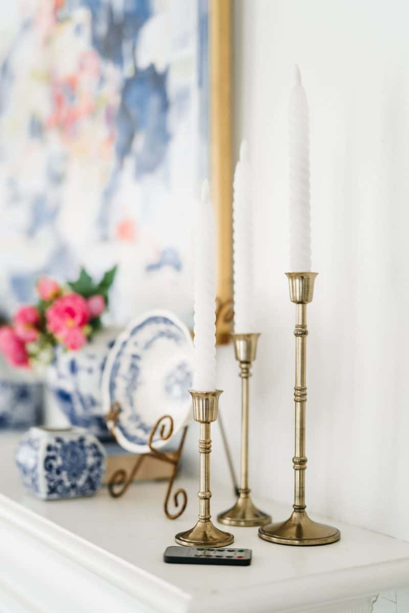 how to mix patterns with blue and white 15 two candles in antique brass holders on a white mantelpiece with a blurred floral painting and vibrant flowers in the background.