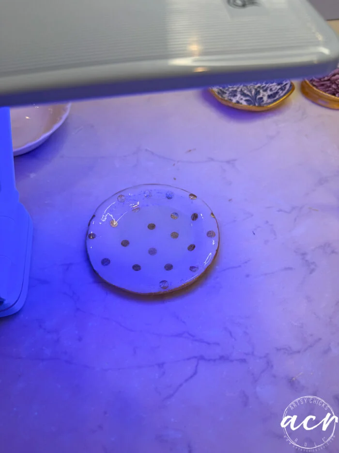 how to use uv resin and decoupage earrings 83 a small round dish with gold polka dots is placed under a uv lamp on a white marble surface, likely curing uv resin. other decorative dishes, possibly for decoupage earrings, are visible in the background, suggesting a creative crafting process.