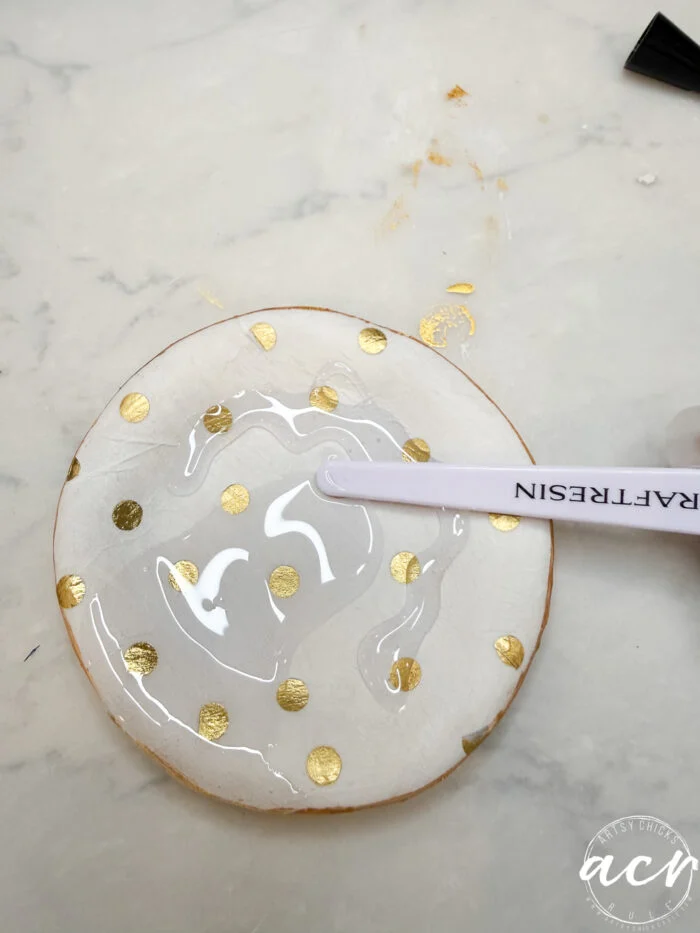 how to use uv resin and decoupage earrings 82 a round white coaster with gold polka dots is being coated with uv resin using a craft stick on a white marble surface, showing how to use resin for a glossy finish.