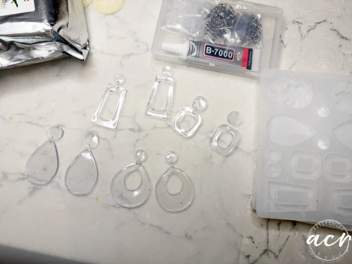 how to use uv resin and decoupage earrings 87 clear resin earring pieces in various shapes are laid out on a marble surface. nearby are a silicone mold, uv resin, b-7000 glue, earring hooks, and a partially visible metallic pouch—perfect for learning how to use decoupage earrings.