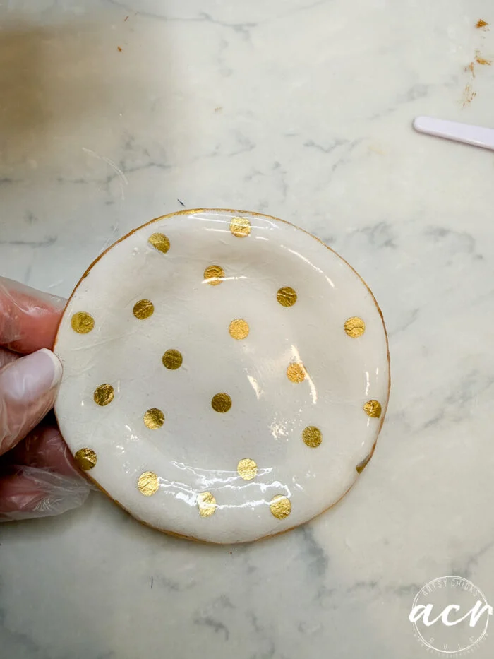 how to use uv resin and decoupage earrings 84 a hand wearing a glove holds a small, round white dish with shiny gold polka dots—perfect for displaying decoupage earrings or curing creations in uv resin—set against a white marble surface.