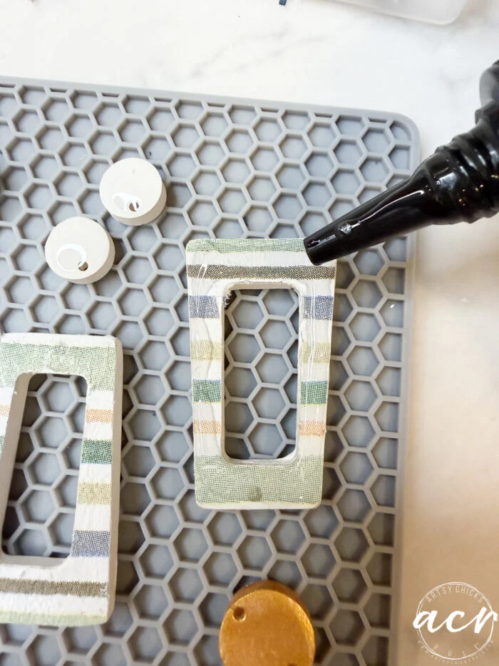 how to use uv resin and decoupage earrings 100 a hand applies glue to a rectangular, striped fabric pendant on a gray hexagonal mat, with circular and round components nearby—perfect for crafting decoupage earrings or learning how to use uv resin.