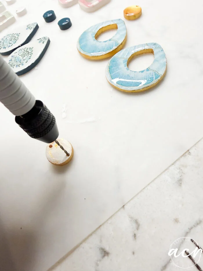 how to use uv resin and decoupage earrings 104 a person uses a power drill to make a hole in a round piece of blue and tan polymer clay, with various uv resin-coated clay earring pieces on a white surface nearby.