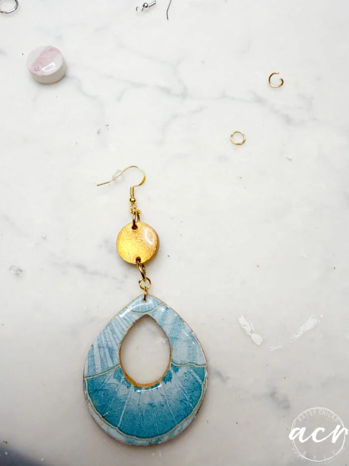 how to use uv resin and decoupage earrings 106 a handmade decoupage earring with a blue teardrop-shaped bottom and a small round gold piece above it rests on a white marble surface, surrounded by a few small jewelry-making components, perfect for learning how to use uv resin.