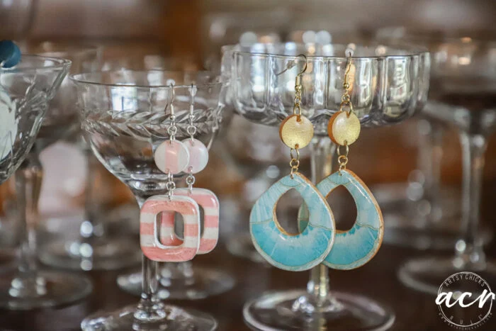 how to use uv resin and decoupage earrings 108 two pairs of colorful decoupage earrings hang from the rims of glass goblets; one pair is pink and white with geometric shapes, the other blue and gold with a teardrop design. the background features more glassware.