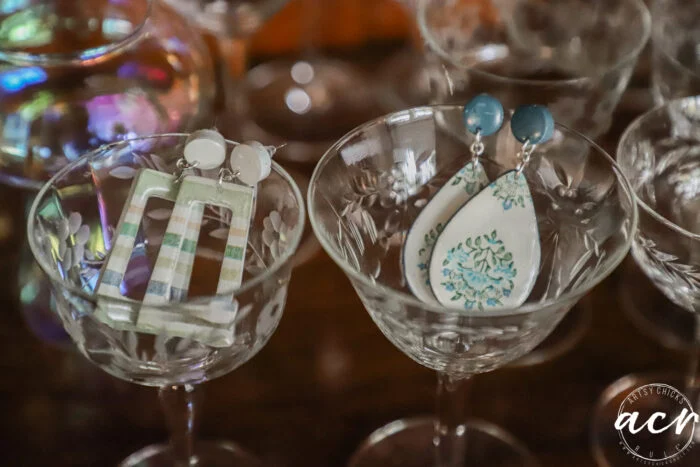 how to use uv resin and decoupage earrings 109 two pairs of decoupage earrings displayed inside vintage etched glass goblets: one pair with green and white stripes, the other with blue and white teardrop shapes. the background features more glassware and soft reflections.