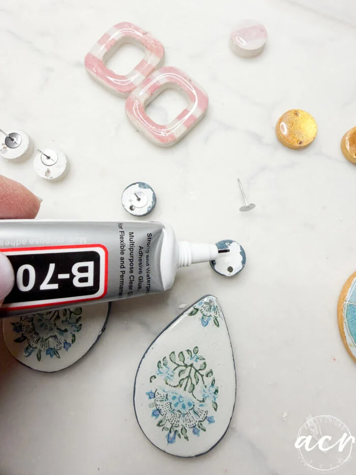 how to use uv resin and decoupage earrings 107 a hand applies b-7000 adhesive to the back of a round earring post; various colorful decoupage earrings and posts are arranged on a white surface, illustrating how to use different materials in jewelry making.