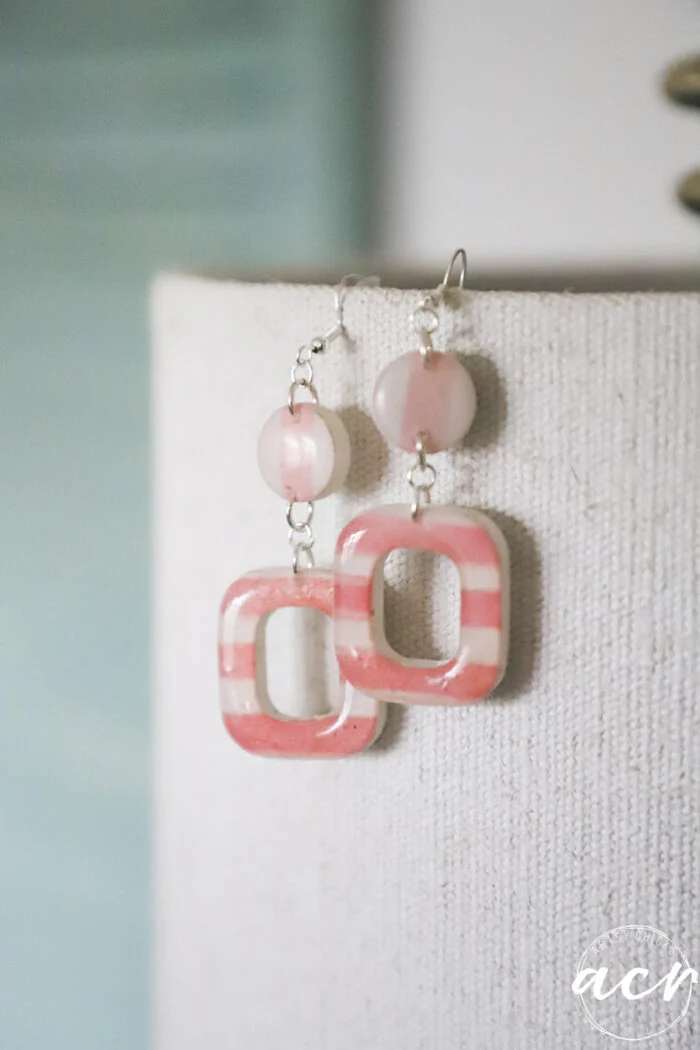 how to use uv resin and decoupage earrings 116 a pair of handmade decoupage earrings with pink round beads and square, pink-striped frames hang from a textured beige fabric background. the soft lighting enhances their artistic style, perfect for learning how to use uv resin in your own creations.