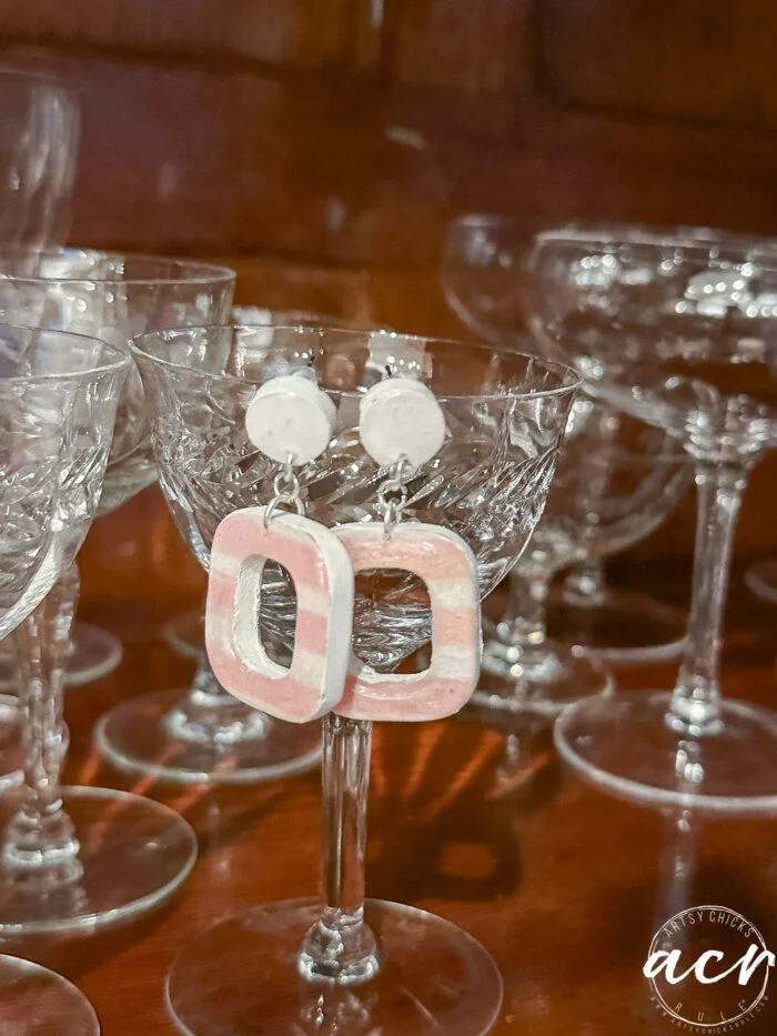 how to use uv resin and decoupage earrings 117 a pair of pink and white square-shaped decoupage earrings with round tops are displayed on the rim of a vintage cut-glass goblet, surrounded by other similar glassware on a wooden surface.