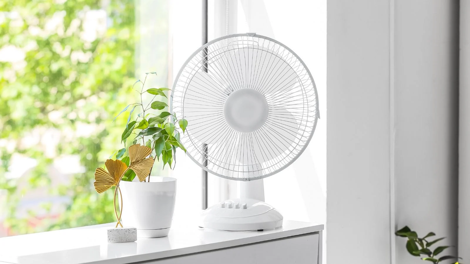 a close-up shot of a small white colored ventilator, placed beside a potted houseplant on a windowsill indoors