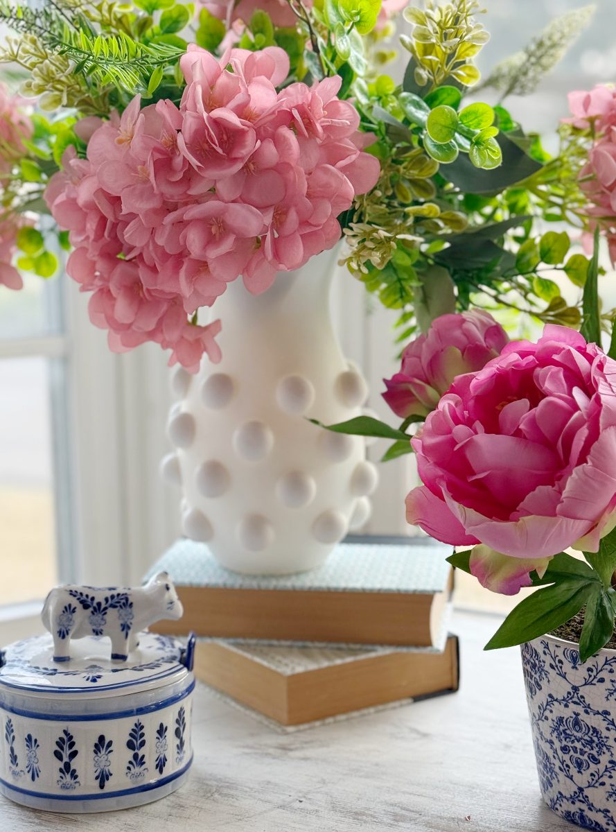 diy hobnail vase that looks like vintage milkglass 32 a white vase with pink flowers sits on two books next to a white pot with blue designs, a small ceramic cow, and a cup with a bright pink peony, all on a bright windowsill.