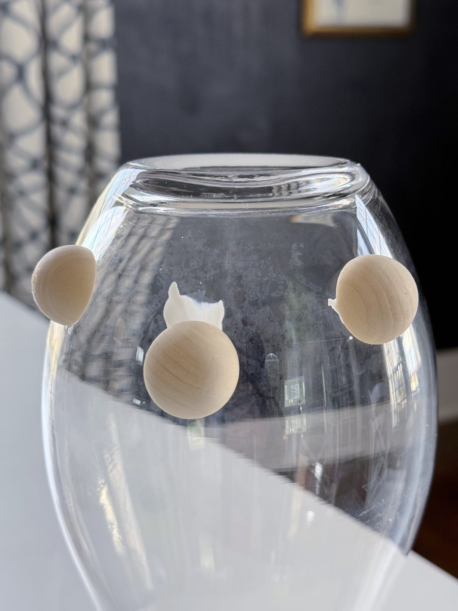 diy hobnail vase that looks like vintage milkglass 35 three wooden beads attached to the outside of an upside-down wine glass, with one bead partially broken. the background shows patterned curtains and part of a picture frame.