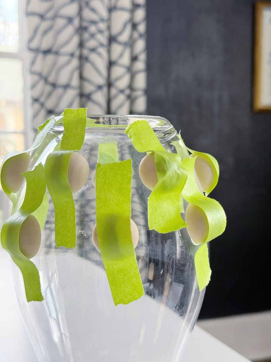 diy hobnail vase that looks like vintage milkglass 36 a clear glass vase has multiple strips of green painter’s tape and round wooden beads attached around its rim. the background features a window with patterned curtains and a dark wall.