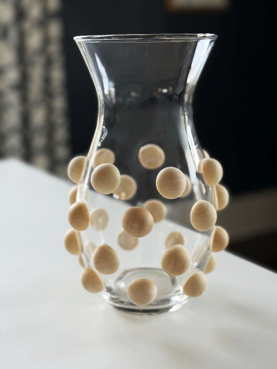 diy hobnail vase that looks like vintage milkglass 38 a clear glass vase with a flared rim, decorated with evenly spaced, round, wooden beads attached to its outer surface. the vase is placed on a white surface with a blurred background.