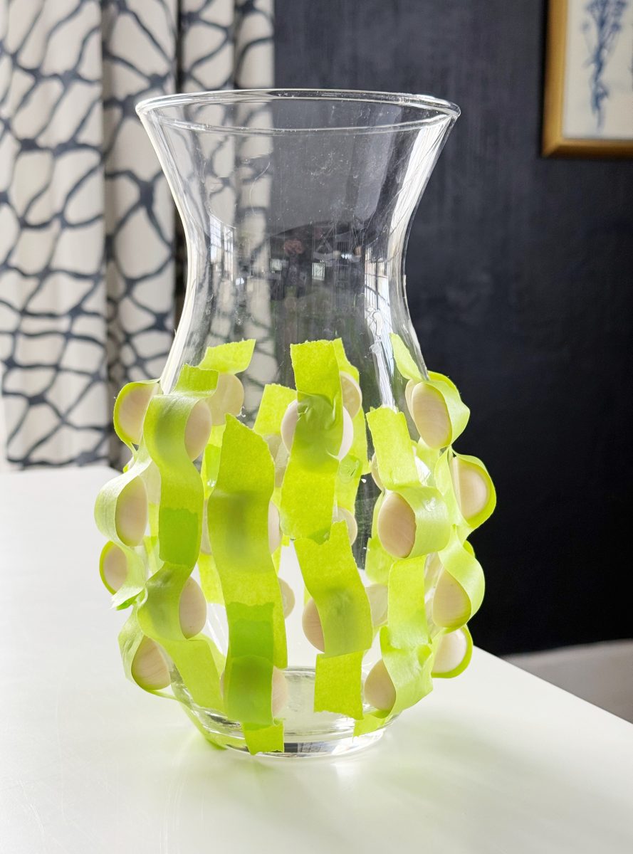 diy hobnail vase that looks like vintage milkglass 37 a clear glass vase decorated with vertical strips of bright green tape that hold small white plastic spoons against its surface, placed on a white table with patterned curtains in the background.