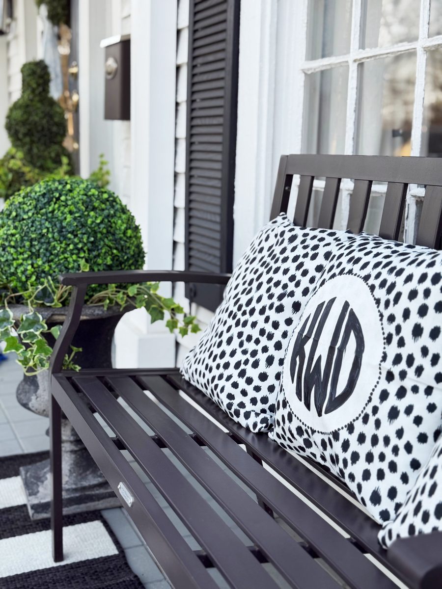 a black metal bench sits on a porch, decorated with spotted black-and-white pillows, one featuring the letters “kwd.” a potted green plant and window with black shutters are nearby.