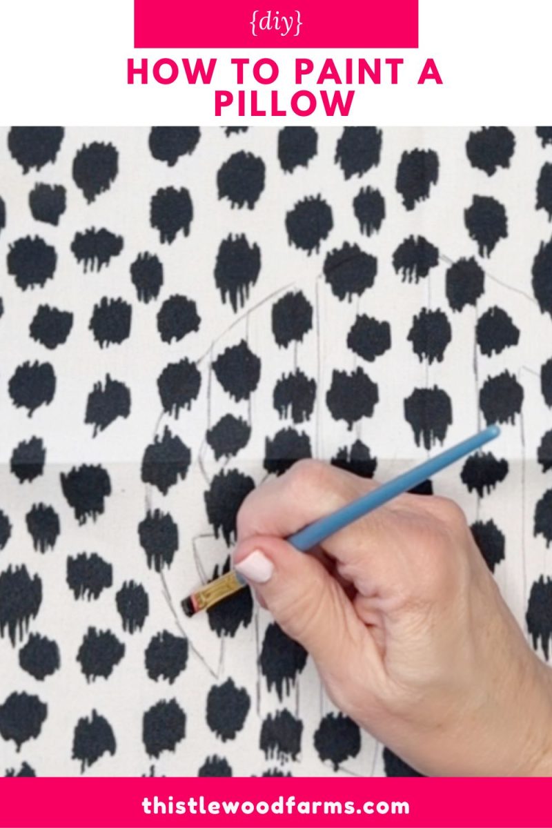 a hand holds a paintbrush, outlining a geometric shape on a white pillow with black abstract spots. the image is labeled "how to paint a pillow" and includes the website thistlewoodfarms.com.