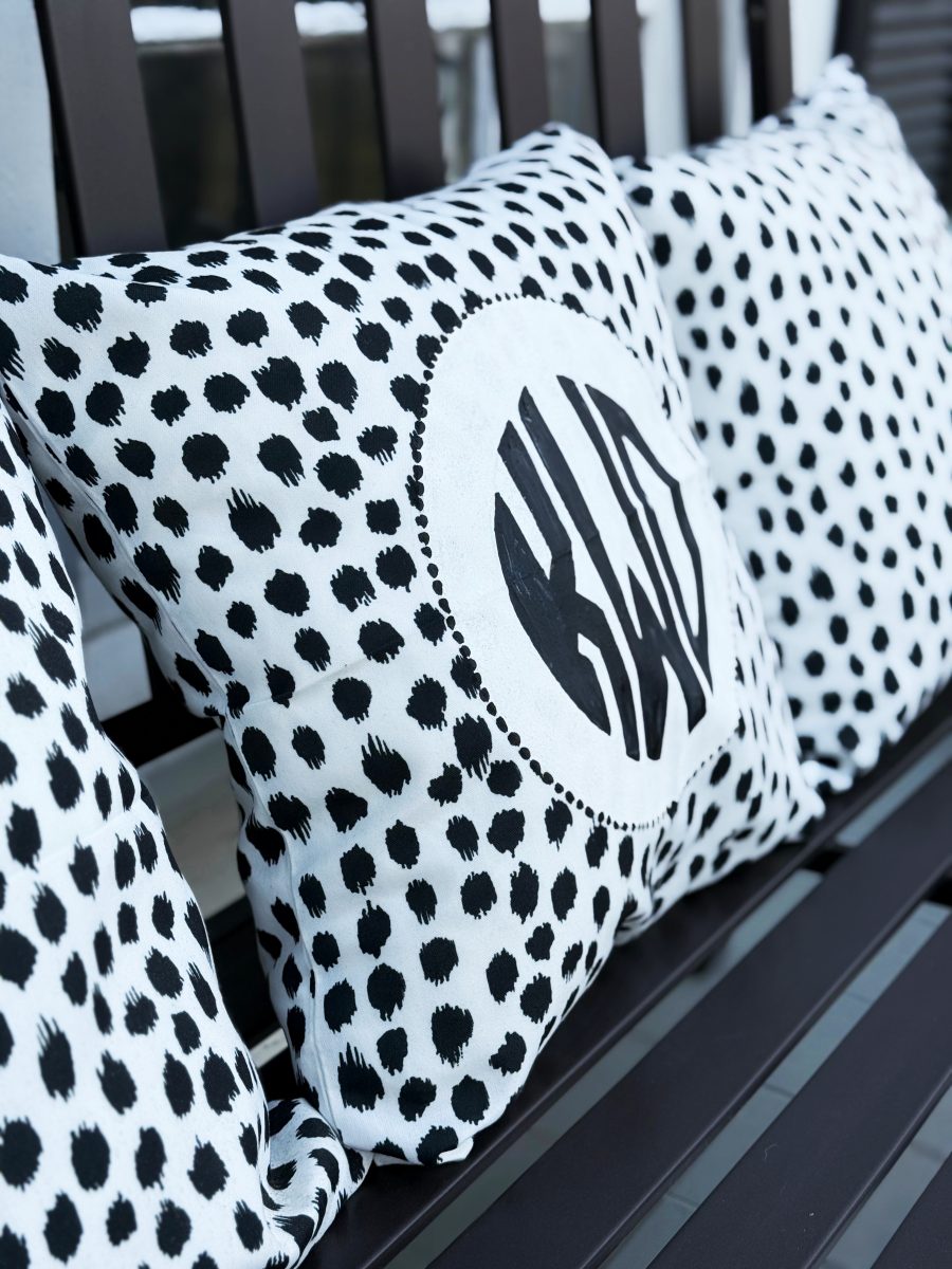 three decorative pillows with black and white polka dot patterns are arranged on a dark bench. the center pillow features a monogram inside a white circle.