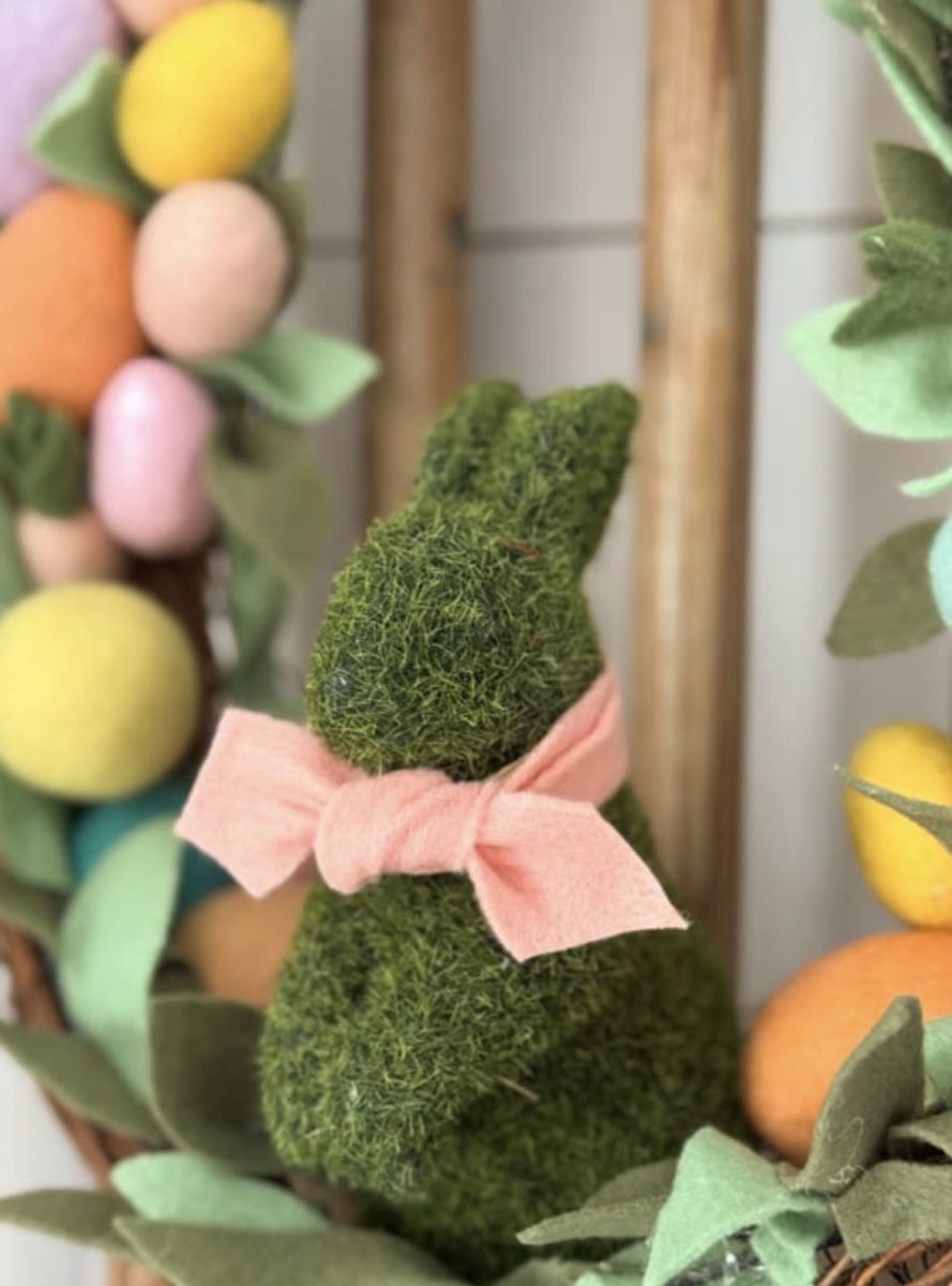 a decorative moss-covered bunny with a pink bow sits among colorful pastel eggs and green felt leaves, creating a festive spring or easter display.