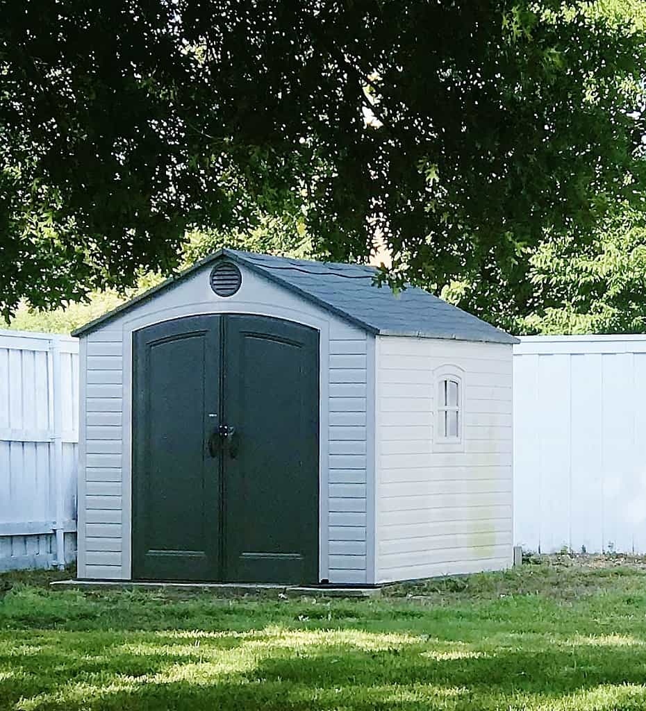 a gray storage shed with gabled roof and double doors, adorned with shed decorations, positioned on a grassy area against a white fence, shaded by leafy trees.