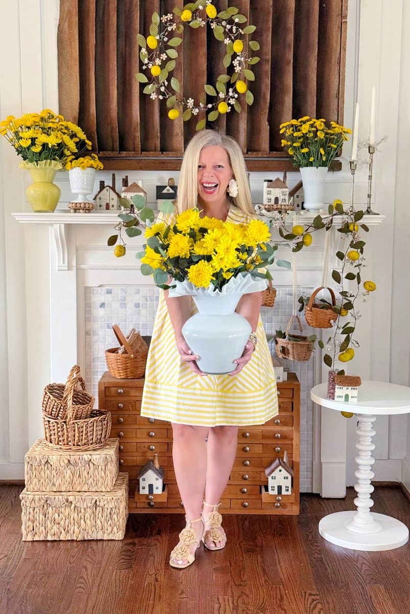a woman in a yellow striped dress stands in front of a decorated fireplace holding a vase of yellow flowers. the mantel is adorned with floral arrangements, baskets, and small decorative houses. she smiles broadly, surrounded by a bright, cheerful setting.