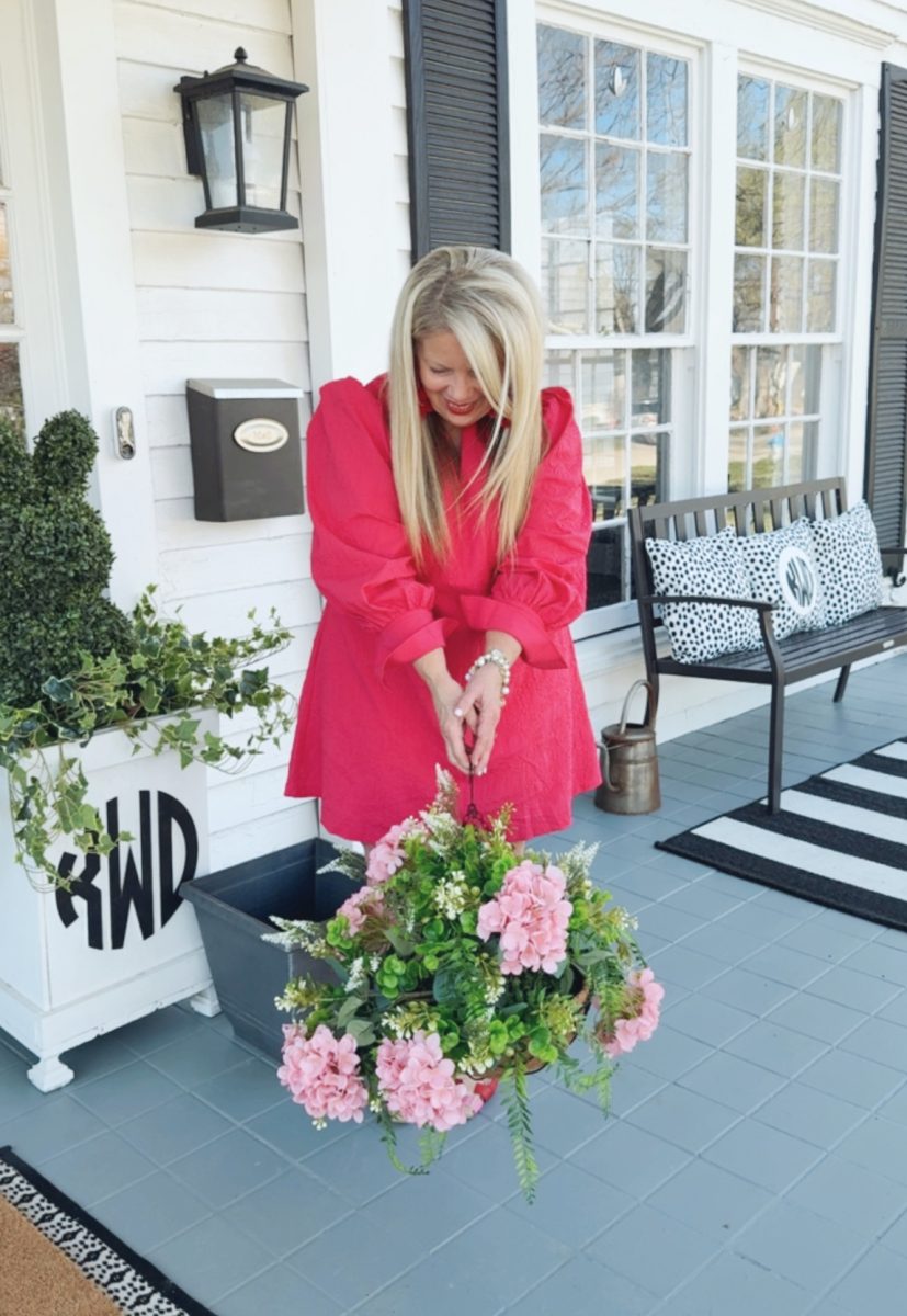 7 easy front porch decor ideas for spring 1 a woman in a bright pink dress stands on a porch, smiling as she holds a basket of pink flowers and greenery. the porch has black-and-white decor, a bench, plants, and striped rugs.