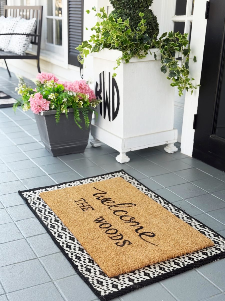 7 easy front porch decor ideas for spring 5 a doormat with the words "welcome the woods" sits on a gray porch near a white planter with greenery and a gray pot of pink flowers. the porch floor has light gray tiles.
