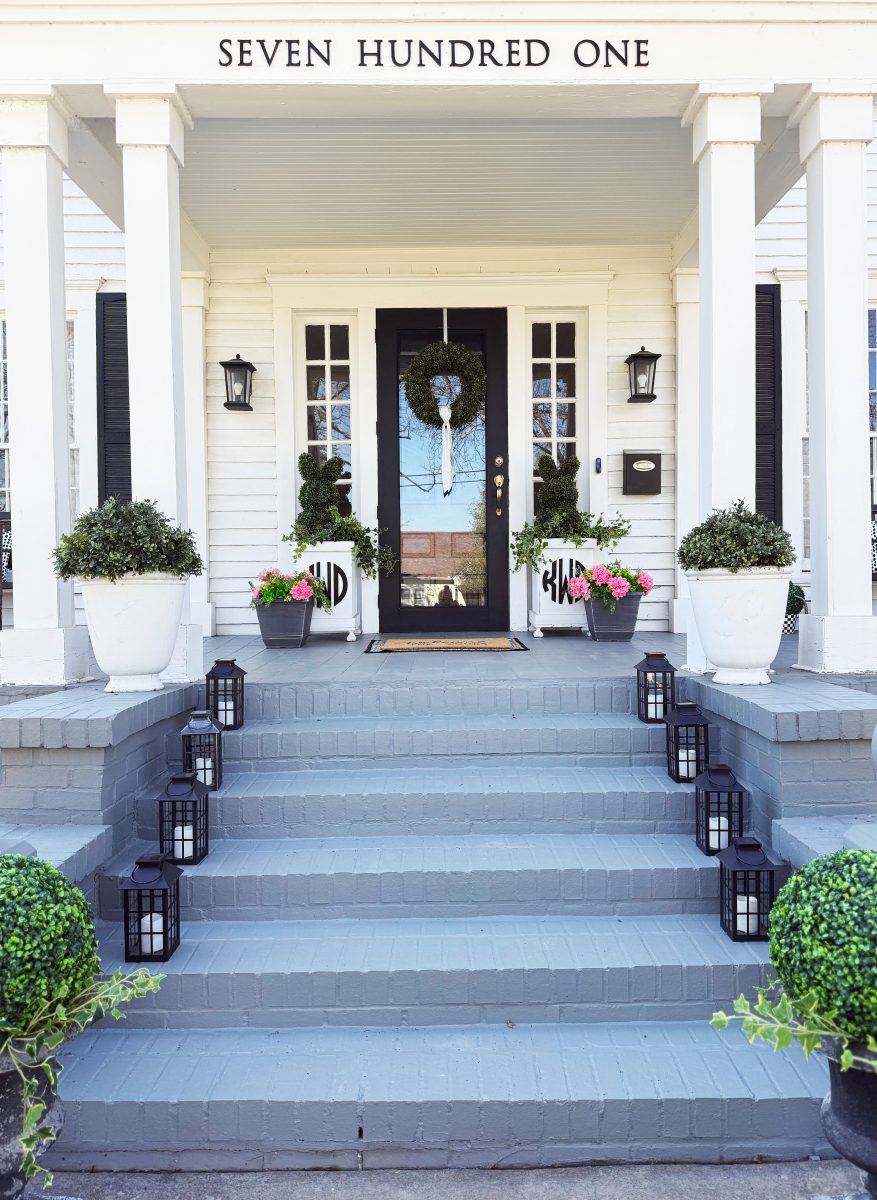 7 easy front porch decor ideas for spring 3 front porch of a white house with gray steps, black door, white columns, potted plants, pink flowers, lanterns lining the steps, and “seven hundred one” written above the entrance.