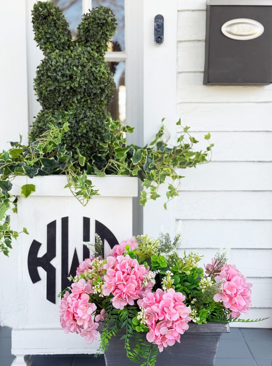 7 easy front porch decor ideas for spring 2 a decorative green topiary shaped like a bunny sits in a white planter with leafy vines. in front, a gray pot holds pink flowers and greenery. the scene is set against a white house with a mailbox and monogram on the planter.