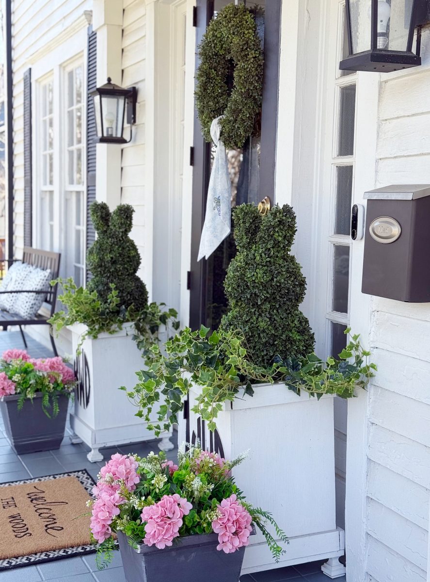 7 easy front porch decor ideas for spring 10 a white porch is decorated with green topiary shaped like bunnies in planters, a wreath on the black door, two pots of pink flowers, and a doormat that says “welcome to the porch.”.